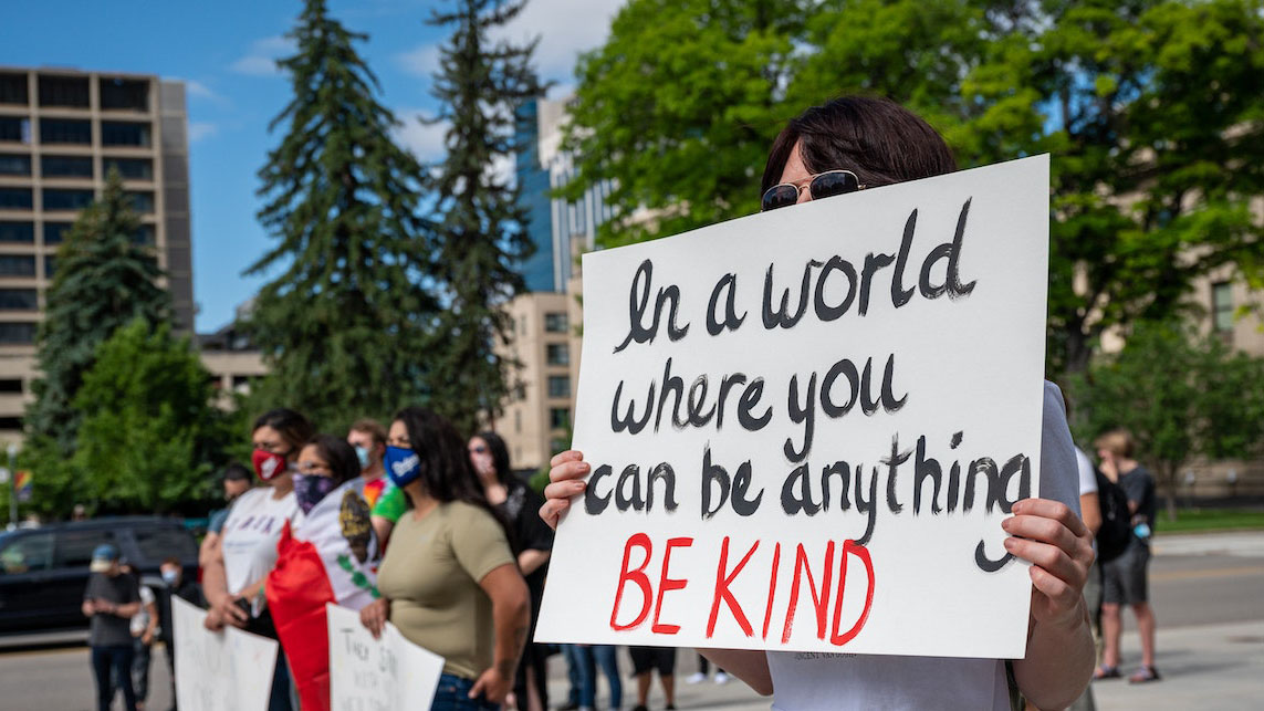 Protestor holding sign that reads, in a world where you can be anything, be kind.